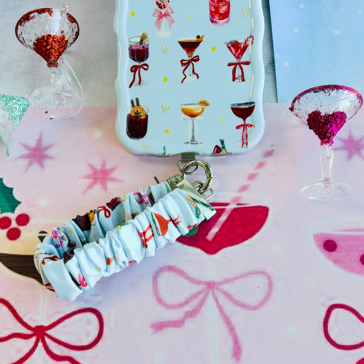 Holiday Spirits, Please? scrunchie phone wrist strap clipped below the matching cocktail print iPhone case on pink cocktail paper