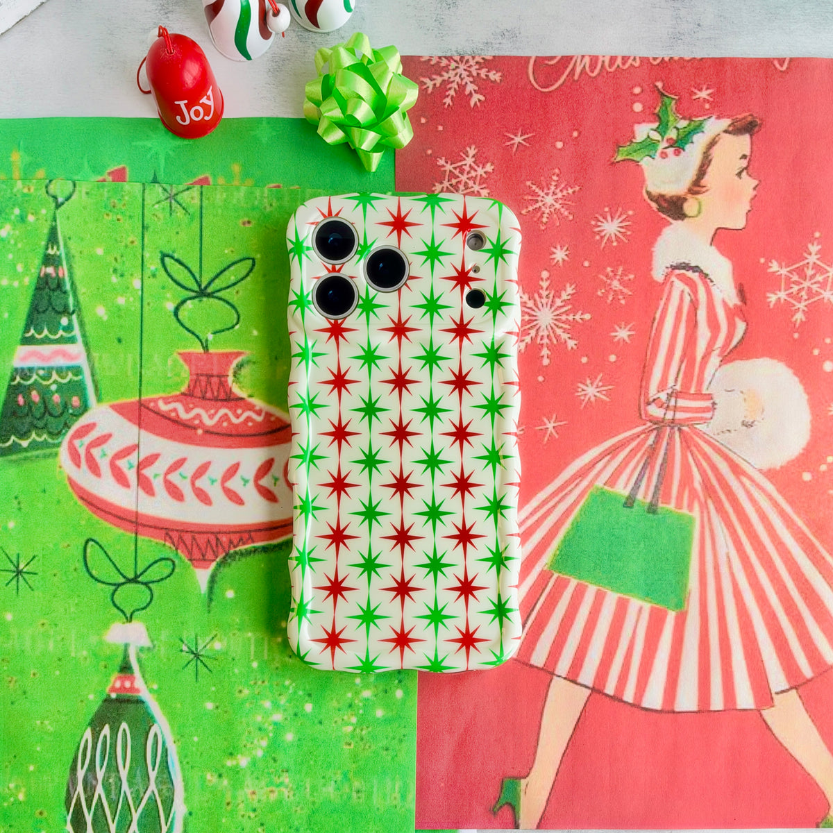Tinsel & Stardust iPhone case lying on retro Christmas paper with a vintage shopping-girl print, a Joy ornament and a lime green gift bow