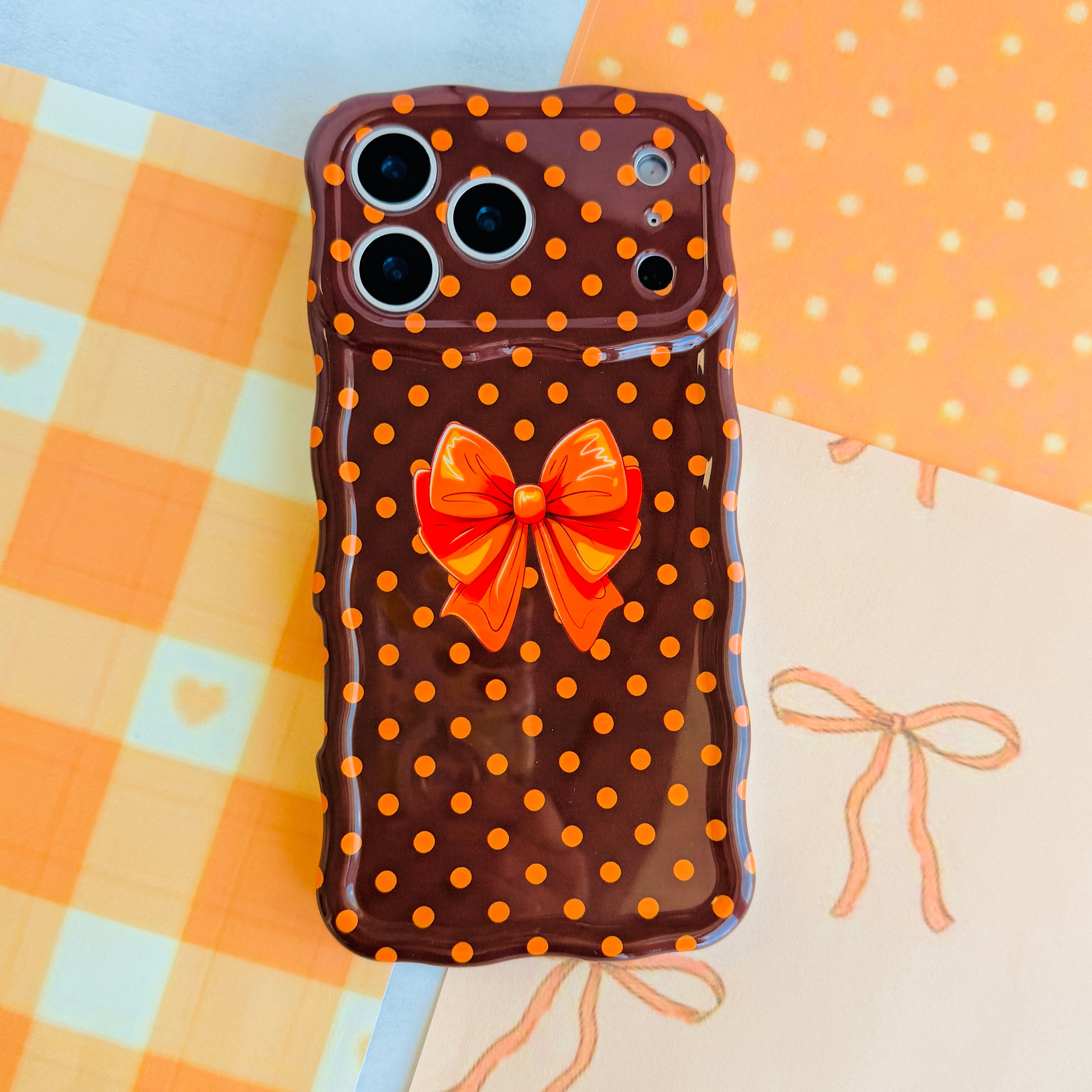 Spotted Looking Cute Wavy Luxe MagSafe iPhone case flat lay on orange gingham, showing the full polka dot print, orange bow, and sculpted wavy edge.