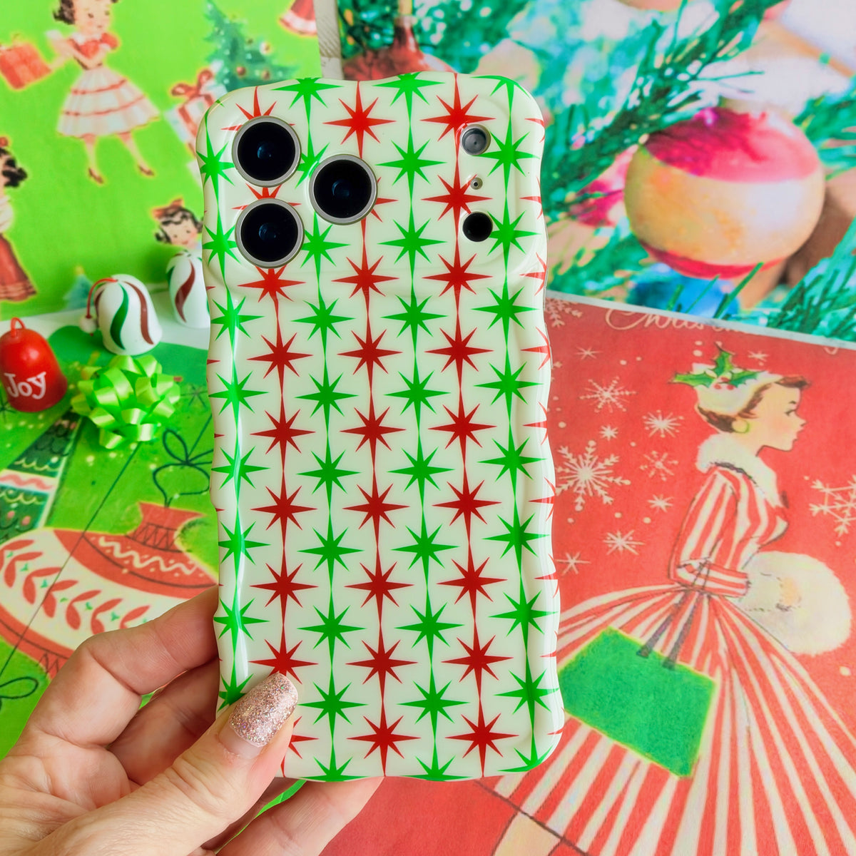 Hand holding the Tinsel & Stardust Wavy Luxe MagSafe iPhone case, red and green starbursts on cream, over vintage 1950s Christmas wrapping paper