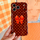 Hand holding the Spotted Looking Cute Wavy Luxe MagSafe iPhone case, showing the chocolate brown base, pumpkin-orange polka dots, and orange bow against an orange gingham and bow backdrop.