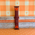 Spotted Looking Cute Apple Watch band single strap half standing against orange gingham, showing the orange bow detail and pumpkin dots on chocolate brown silicone.