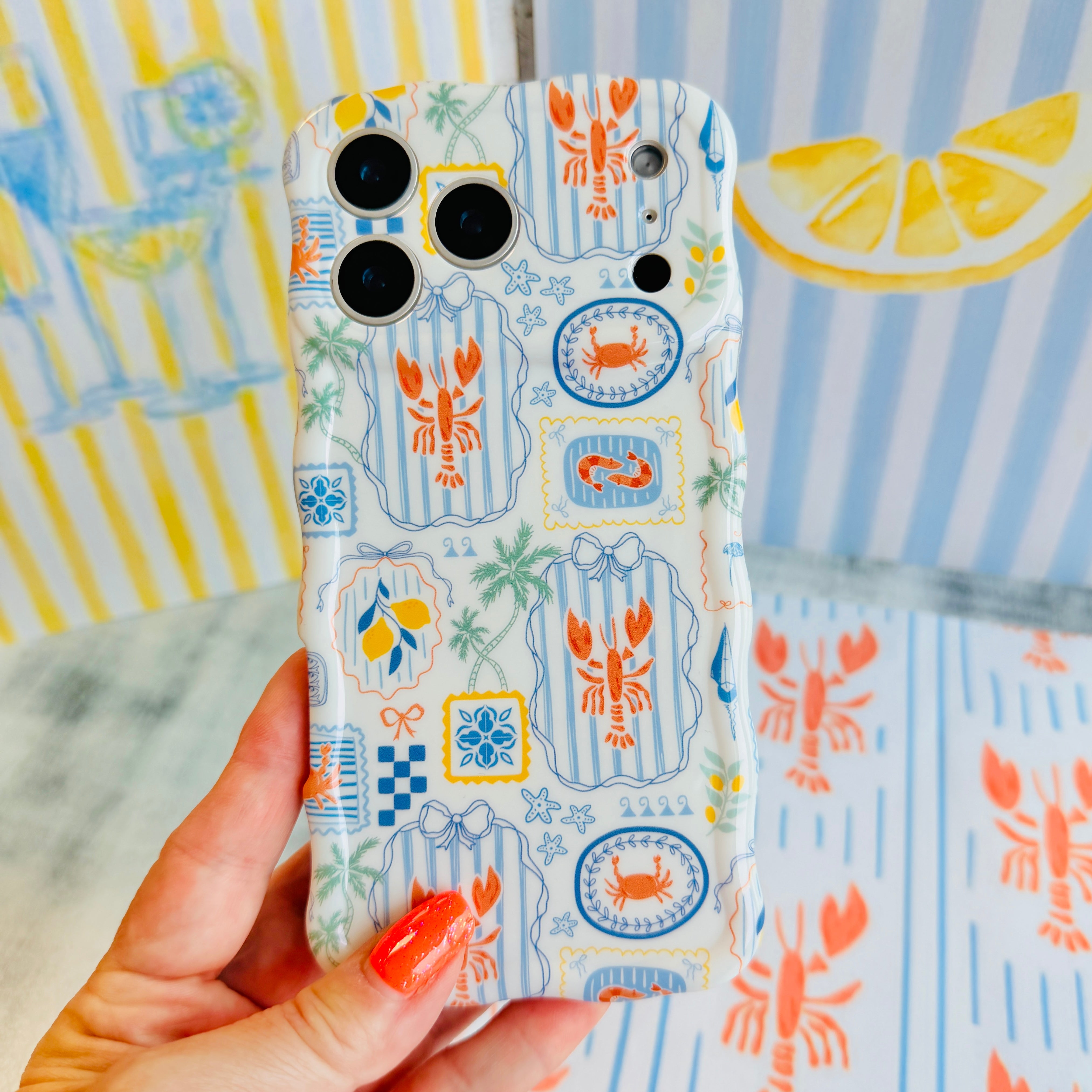 Hand holding the Coastal Catch Wavy Luxe MagSafe iPhone case with lobsters, crabs and blue tiles on a striped background