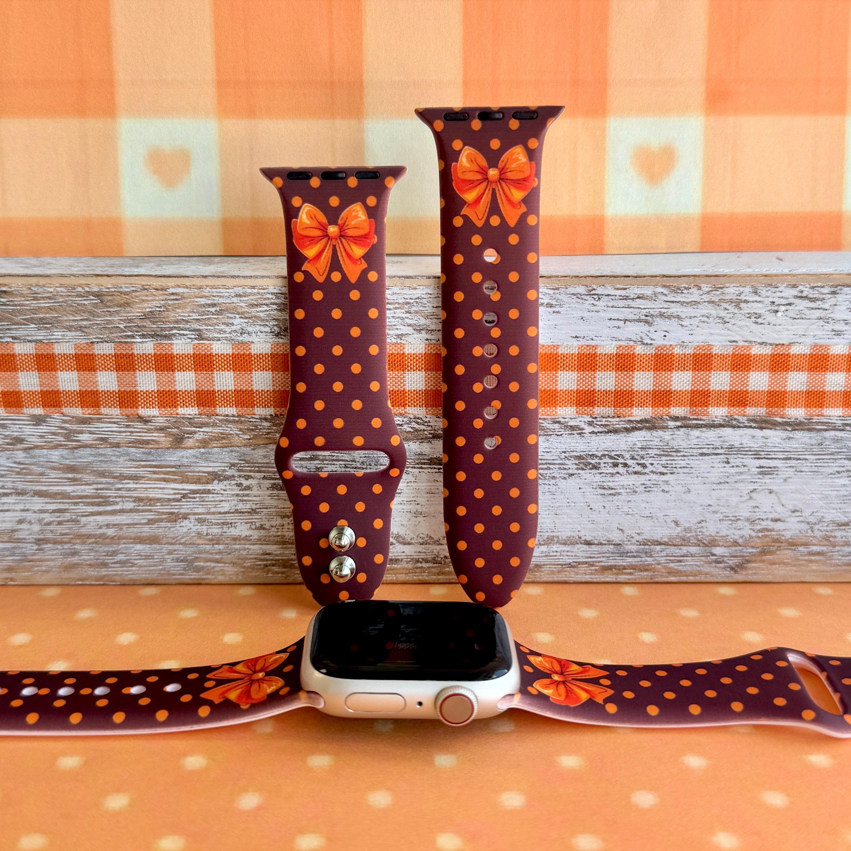 Spotted Looking Cute silicone Apple Watch band shown with an Apple Watch, both strap halves standing upright to display the chocolate brown base, pumpkin-orange polka dots, and orange bow.