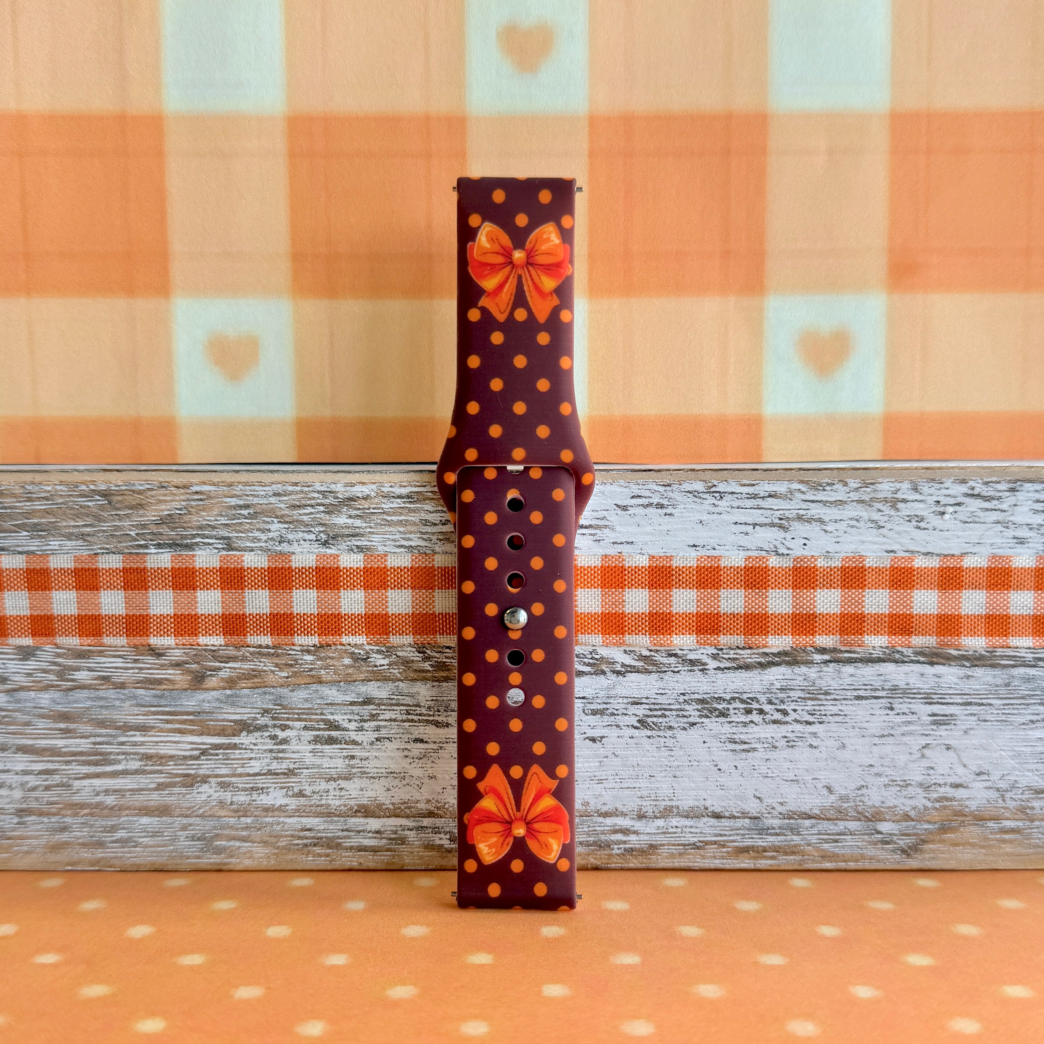 Spotted Looking Cute Samsung Galaxy Watch band full-length strap half on weathered wood and orange gingham, showing the bow detail and polka dot print end to end.