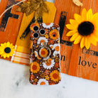 Harvest Sunflowers MagSafe grip ring attached to the matching Harvest Sunflowers iPhone case, styled against an orange barn wood fall sign with real sunflowers and orange plaid.