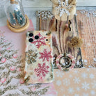 Champagne Snowfall grip ring lying beside the matching pink and gold snowflake iPhone case on whitewashed wood with a wooden sleigh