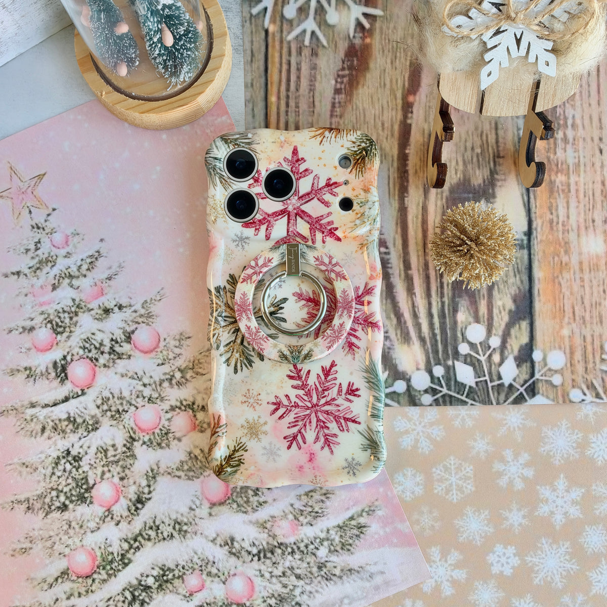 Champagne Snowfall iPhone case with the matching snowflake MagSafe grip ring snapped on, with a gold glitter pom and wooden sleigh