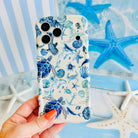 Hand holding the Blue Lagoon Wavy Luxe MagSafe iPhone case in blue watercolor sea turtles and shells on a coastal background