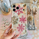 Hand holding the Champagne Snowfall Wavy Luxe MagSafe iPhone case, pink and gold glitter snowflakes, over a pink snowy-pine backdrop