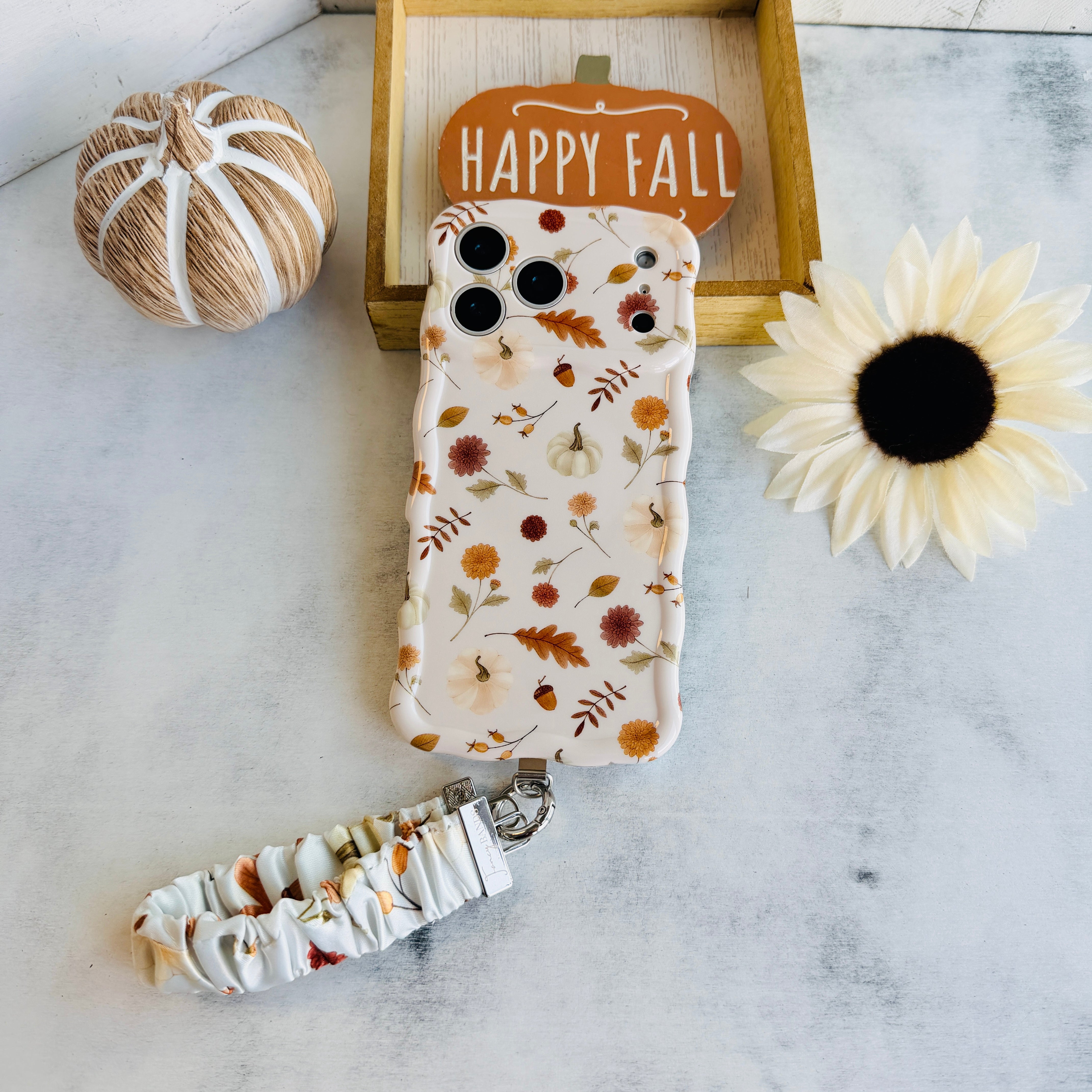 Fall Floral Bliss scrunchie phone wrist strap attached to the matching Fall Floral Bliss iPhone case, photographed with a Happy Fall pumpkin sign, a twine pumpkin, and a cream sunflower.