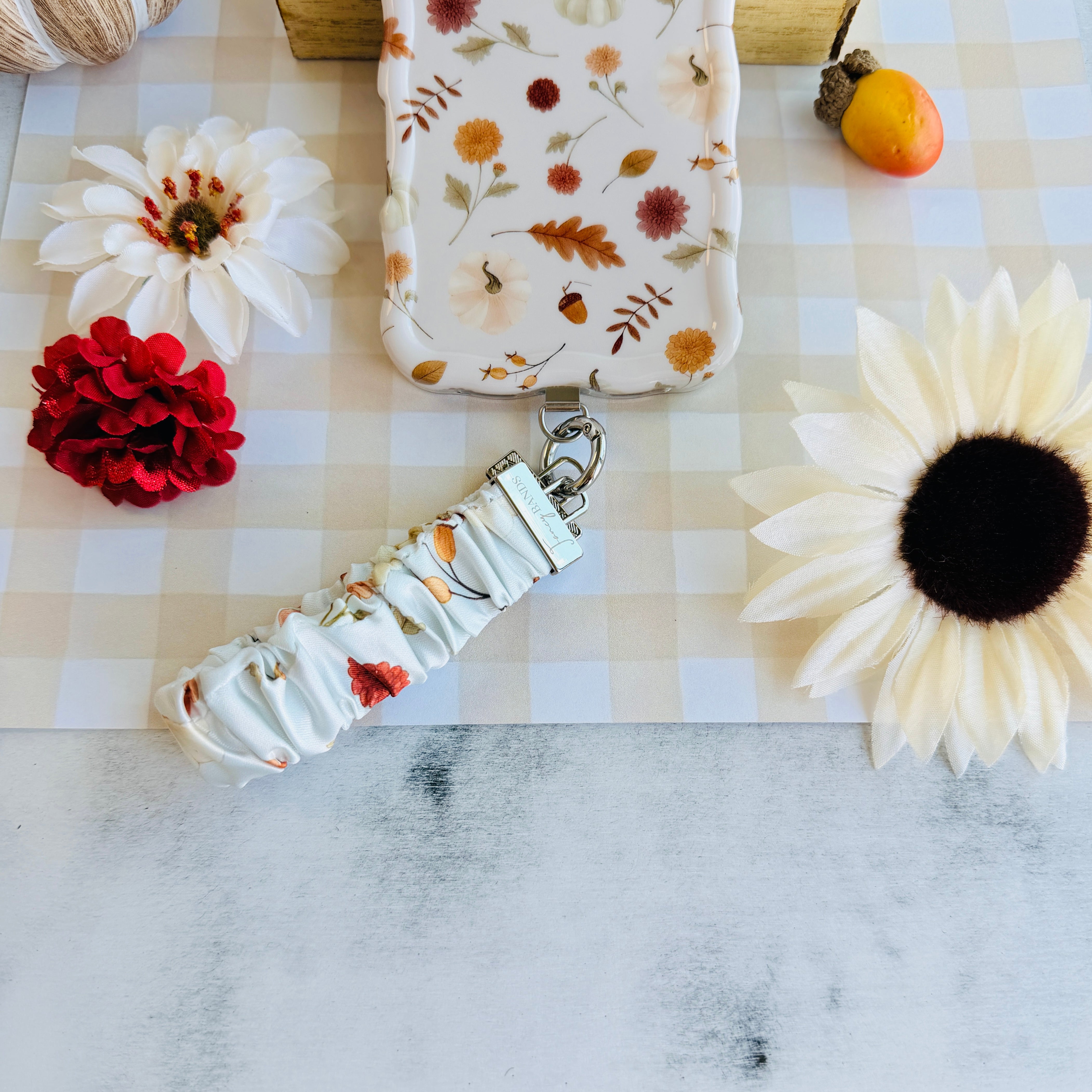 Fall Floral Bliss scrunchie phone wrist strap clipped to the matching Fall Floral Bliss Wavy Luxe iPhone case, styled on cream gingham with a white dahlia, a cream sunflower, a small orange pumpkin, and an acorn.