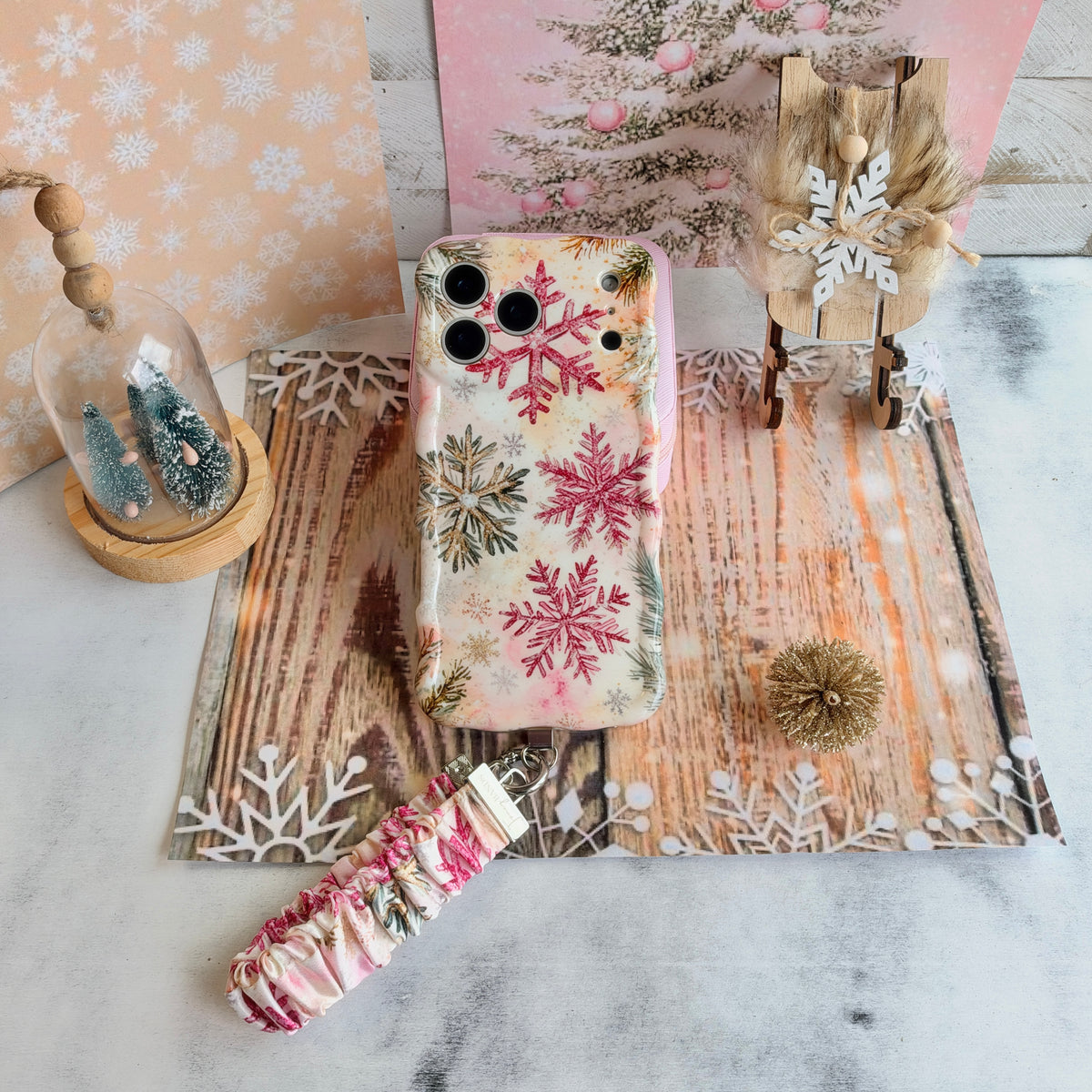 Champagne Snowfall iPhone case and its pink and gold snowflake scrunchie wrist strap beside a cloche of bottle-brush trees