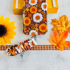 Harvest Sunflowers scrunchie phone wrist strap clipped to the matching Harvest Sunflowers Wavy Luxe iPhone case, styled on orange gingham ribbon with a sunflower, autumn maple leaves, and a yellow wooden crate.