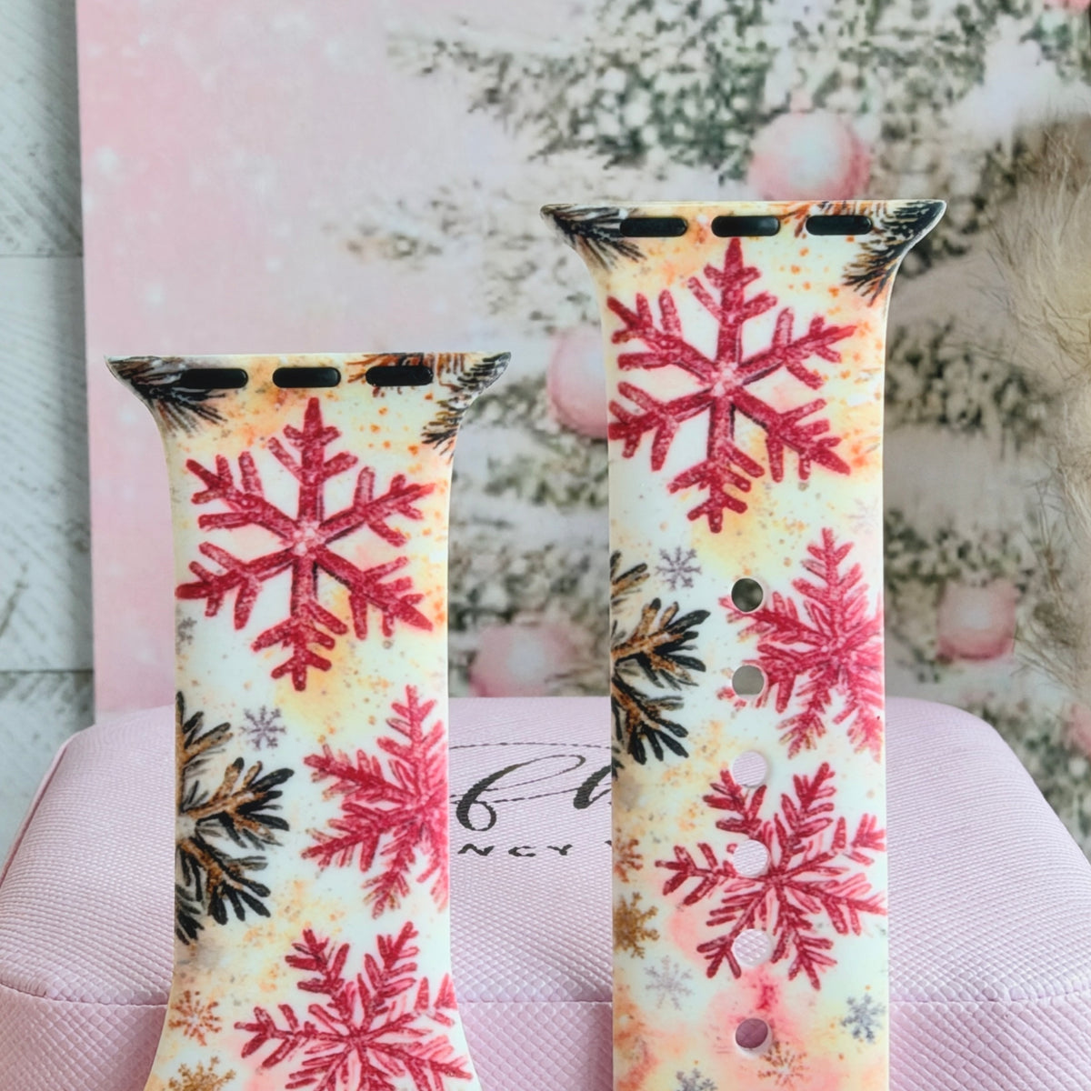 Close-up of both Champagne Snowfall Apple Watch band straps with glittery pink snowflakes and pine sprigs on a peach watercolor ground