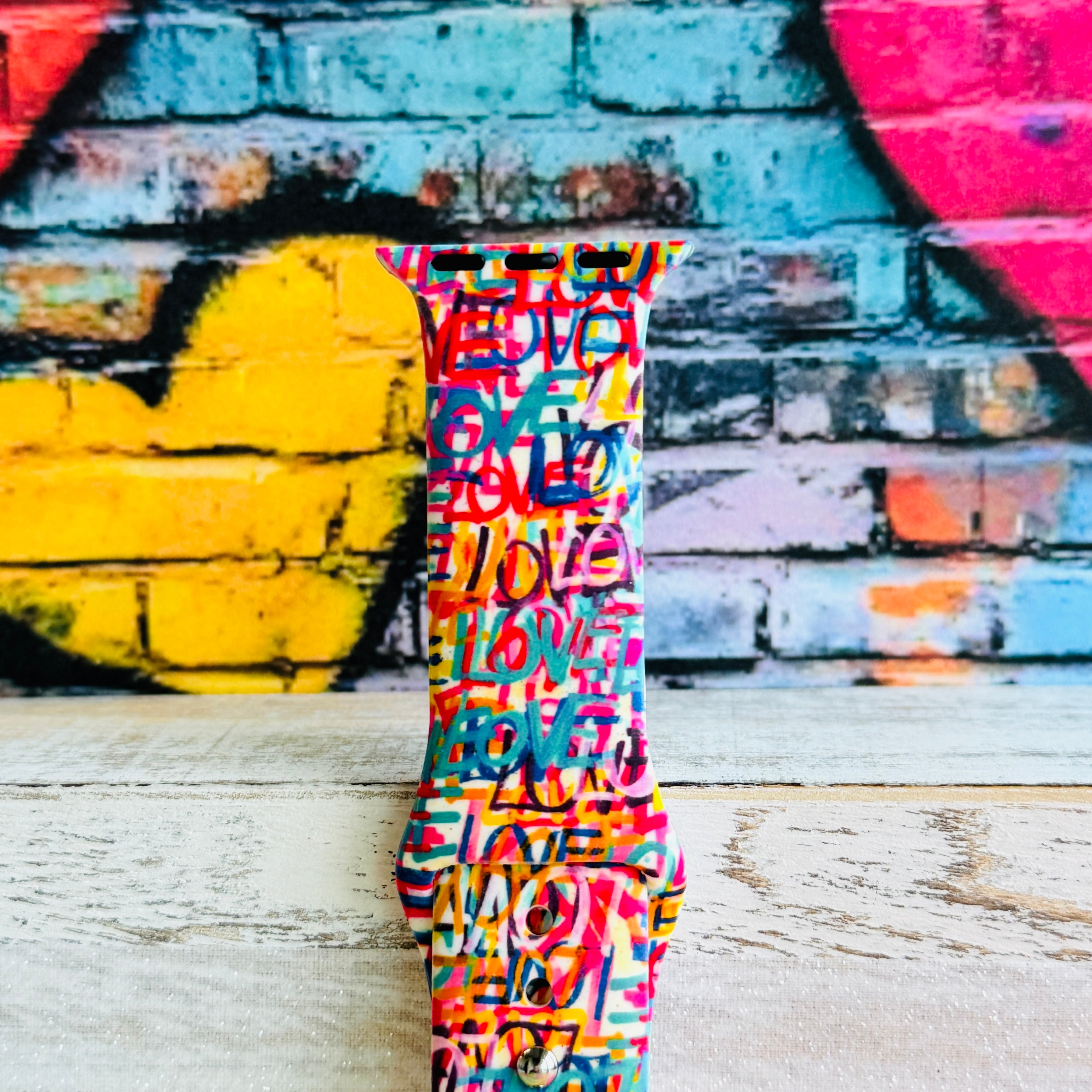 Love Riot Apple Watch band with colorful graffiti "LOVE" text in red, pink, yellow, blue, and turquoise with street art backdrop
