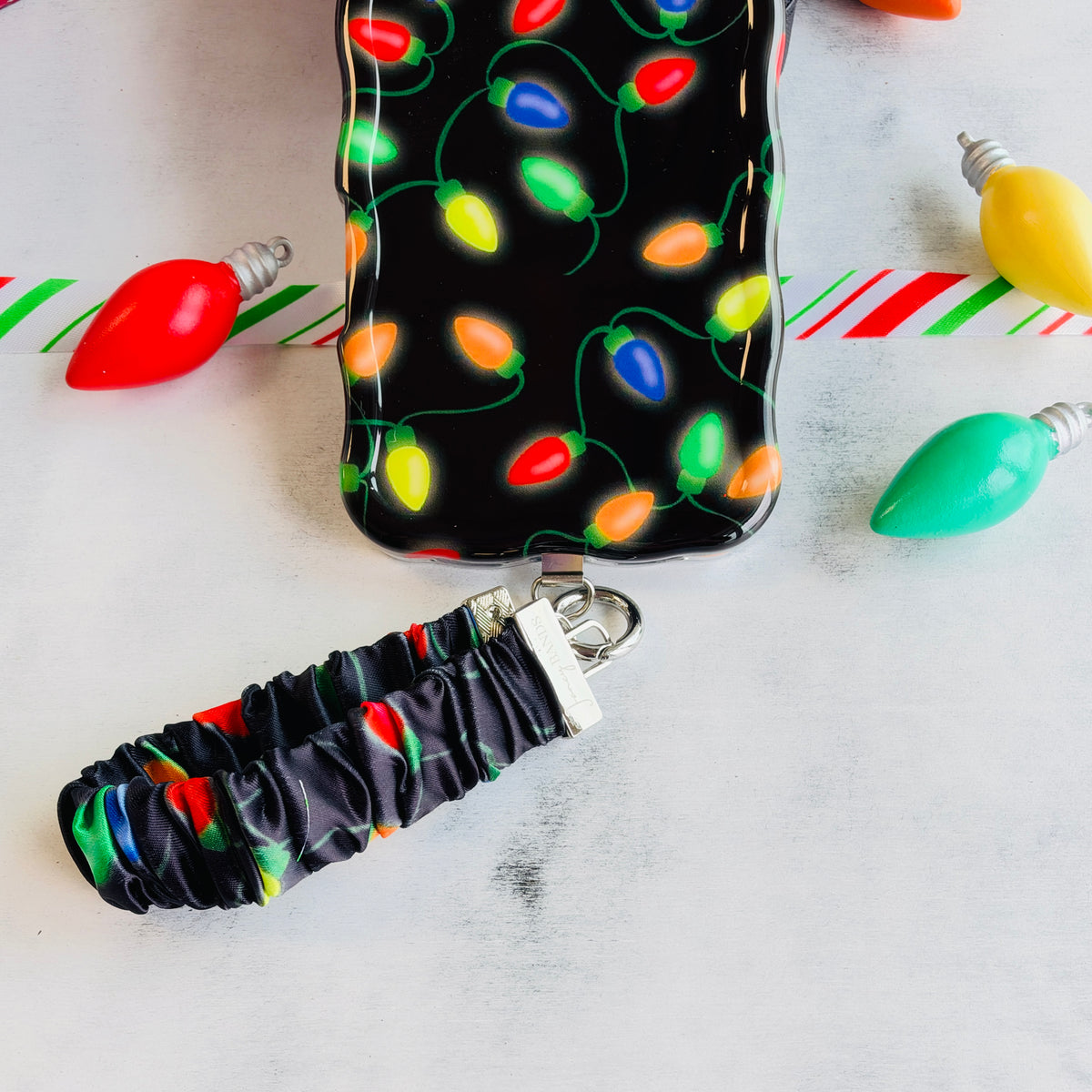 Christmas Lights scrunchie phone wrist strap clipped below the matching iPhone case, with oversized red, yellow and green bulb ornaments