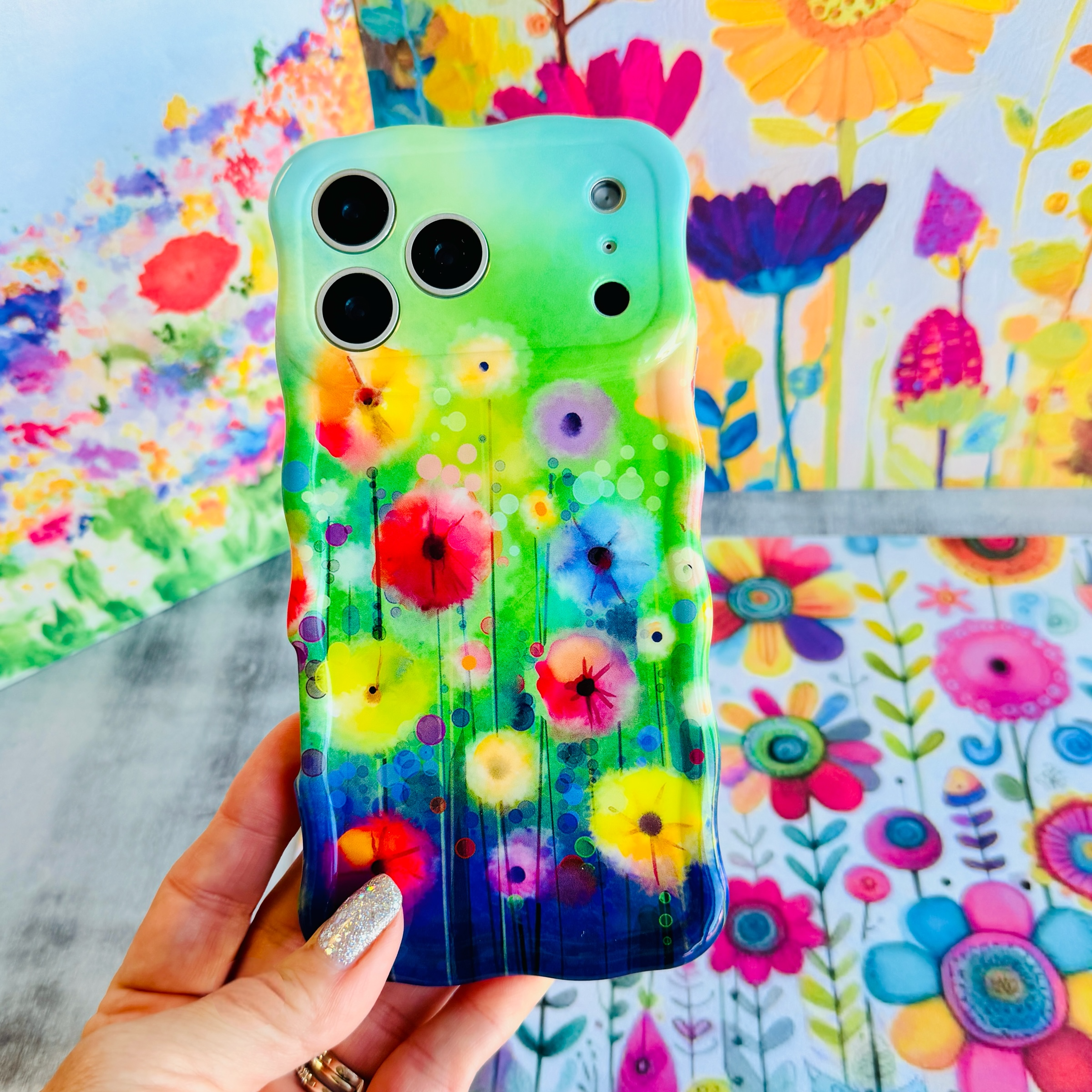 Hand holding the Petal Princess Wavy Luxe MagSafe iPhone case with painted rainbow wildflowers on a green and blue wash