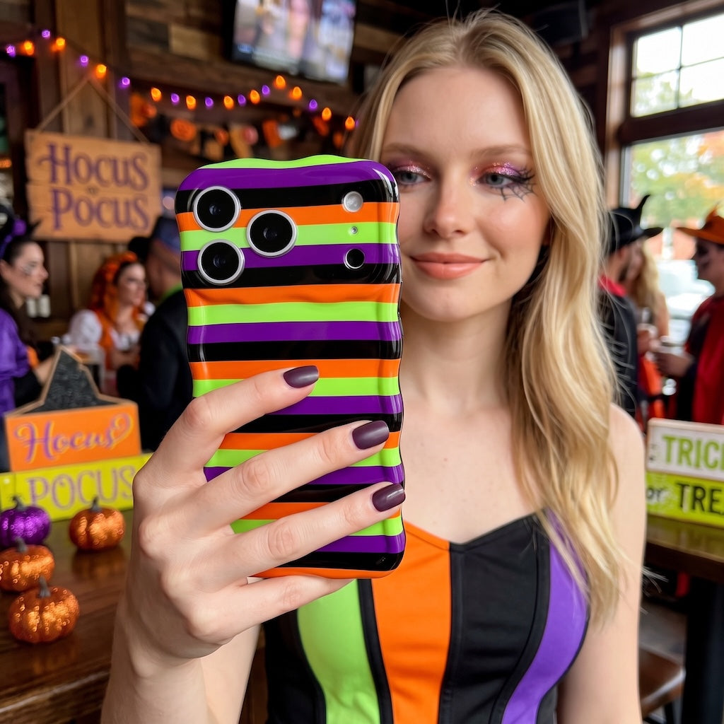 Woman holding the striped Main Witch Energy Wavy Luxe MagSafe iPhone case at a Halloween party beside a Hocus Pocus sign.
