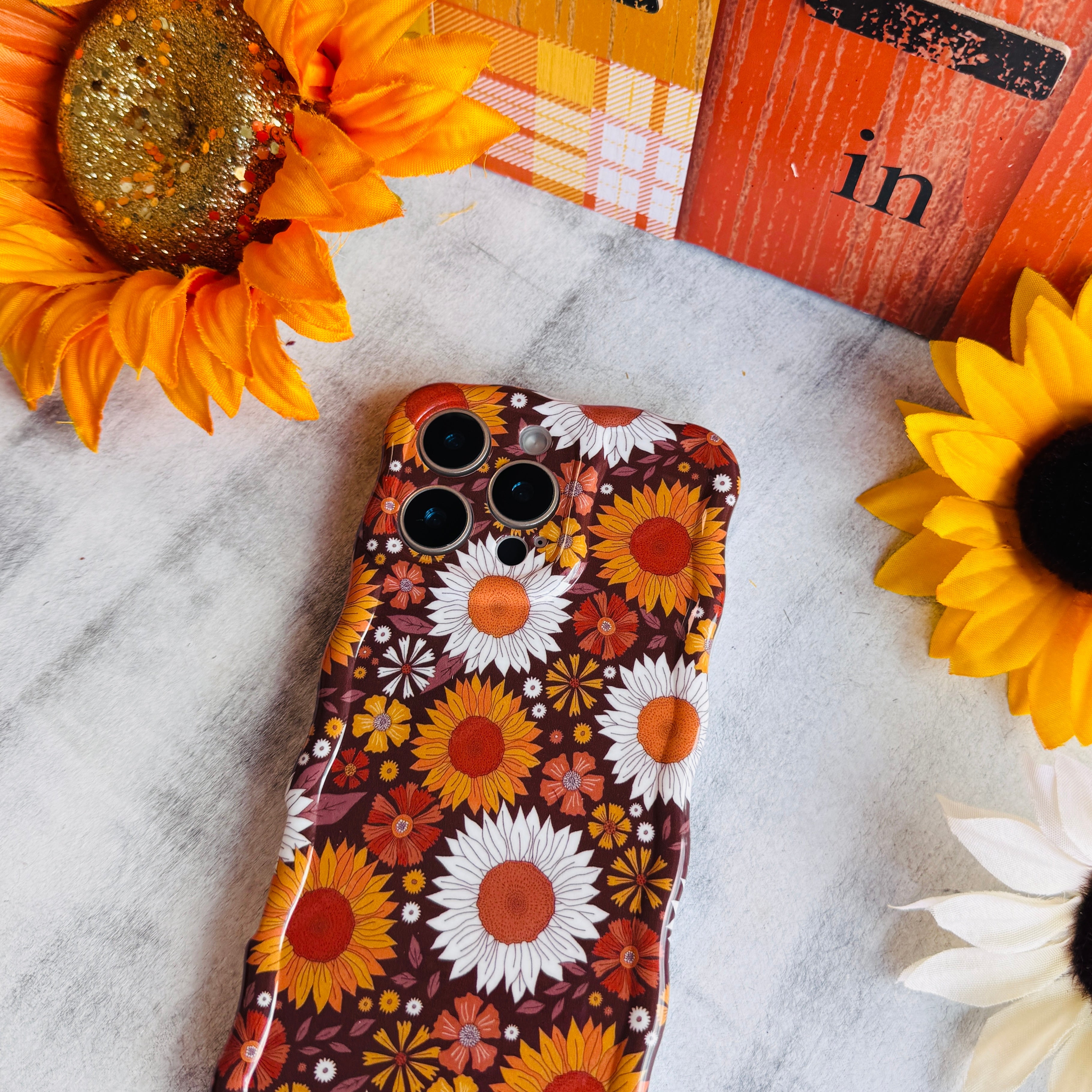 Harvest Sunflowers Wavy Luxe MagSafe iPhone case with golden sunflowers and white daisies on chocolate brown, styled with fall decor
