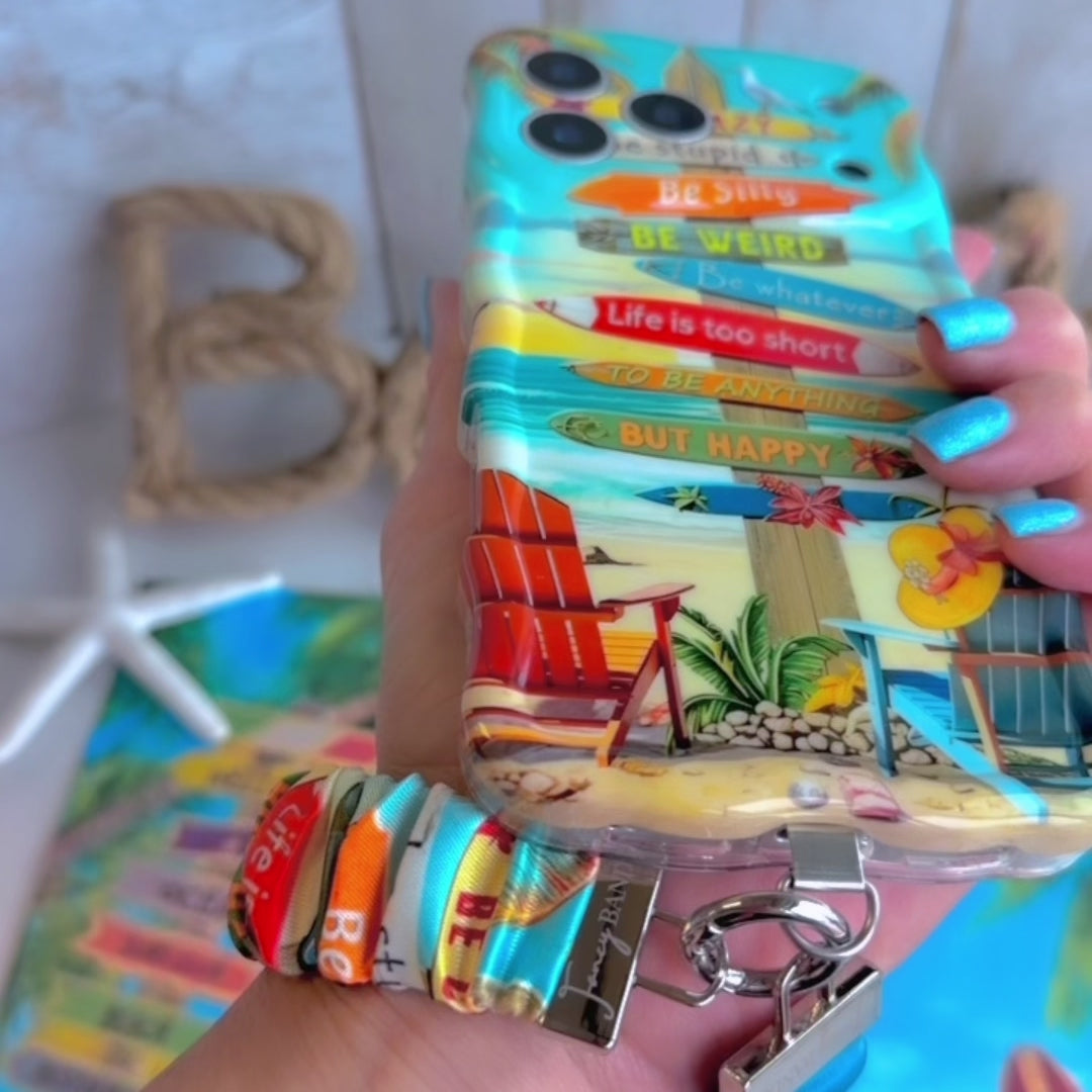 A colorful Seaside State Of Mind scrunchie phone wrist strap in a vintage beach print with Adirondack chairs, surfboard signposts, and palm trees, clipped to the matching Seaside State Of Mind Wavy Luxe iPhone case, styled on a tropical beach background with starfish and shells.
