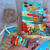 A colorful Seaside State Of Mind scrunchie phone wrist strap in a vintage beach print with Adirondack chairs, surfboard signposts, and palm trees, clipped to the matching Seaside State Of Mind Wavy Luxe iPhone case, styled on a tropical beach background with starfish and shells.