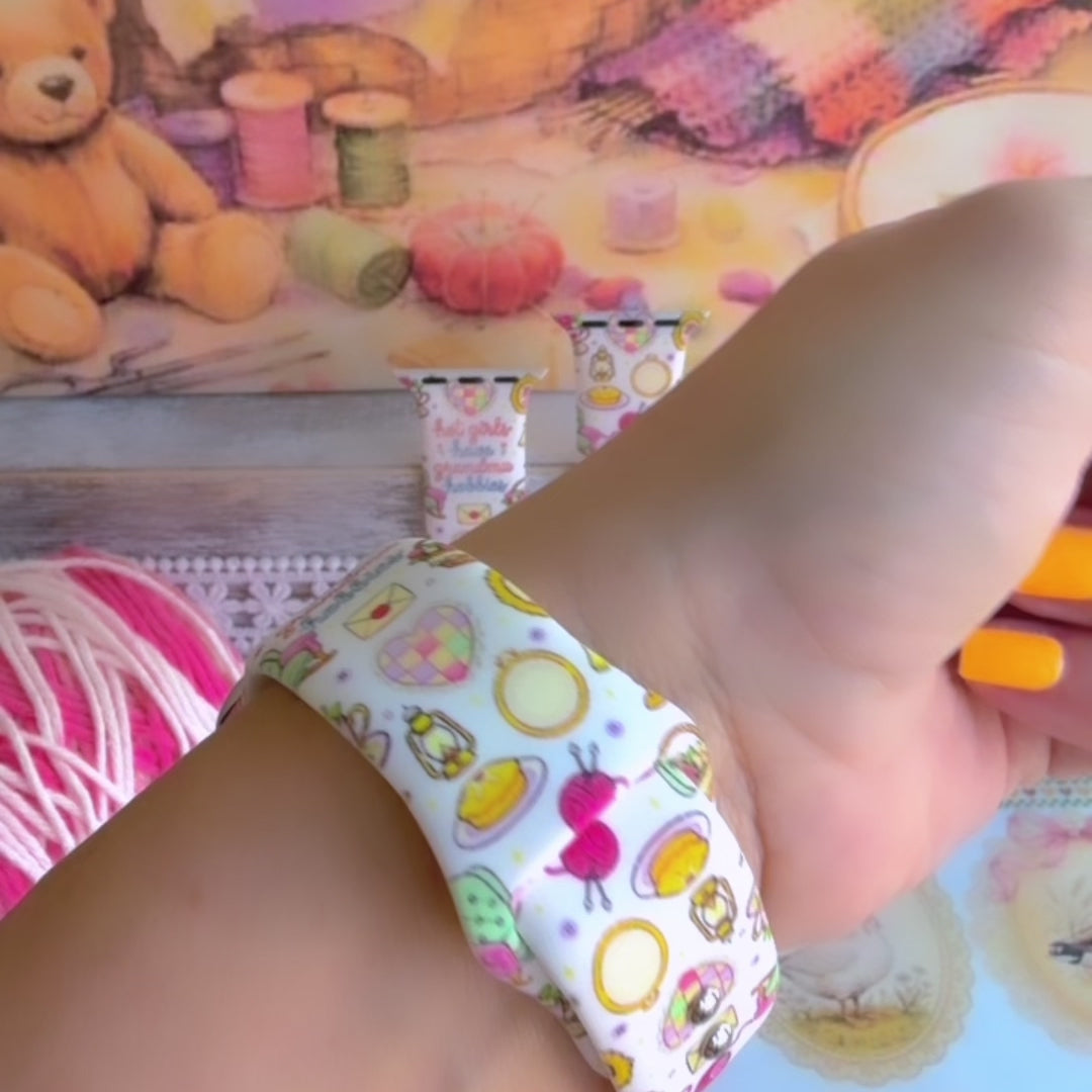 It's Giving Grandma Hobbies silicone Apple Watch band with "hot girls have grandma hobbies" script, patchwork hearts, yarn balls, knitting needles, embroidery hoops, lanterns, pies, and teacups on a cream base, shown displayed and worn on Apple Watch against a cottagecore craft backdrop.