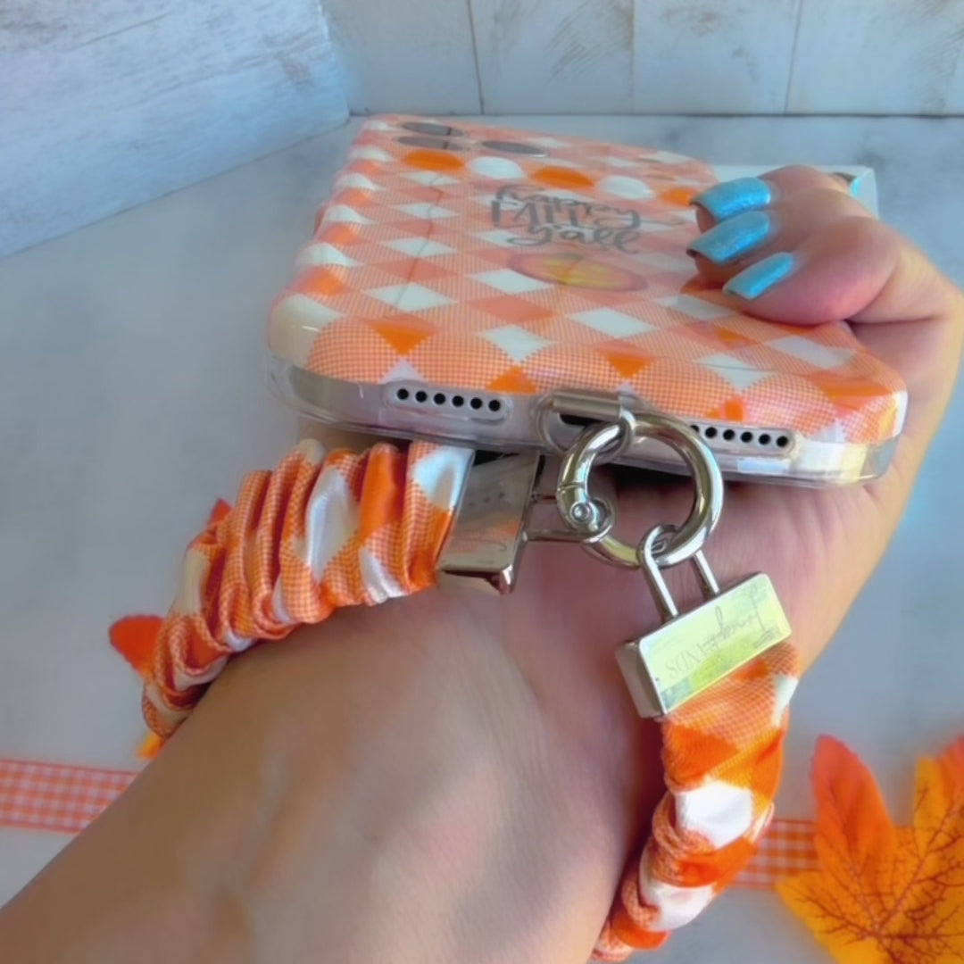 An orange gingham Happy Fall Y'all scrunchie phone wrist strap with a jack-o'-lantern and 'happy fall y'all' print, clipped to the matching Happy Fall Y'all Wavy Luxe iPhone case, styled with autumn maple leaves.