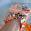 An orange gingham Happy Fall Y'all scrunchie phone wrist strap with a jack-o'-lantern and 'happy fall y'all' print, clipped to the matching Happy Fall Y'all Wavy Luxe iPhone case, styled with autumn maple leaves.