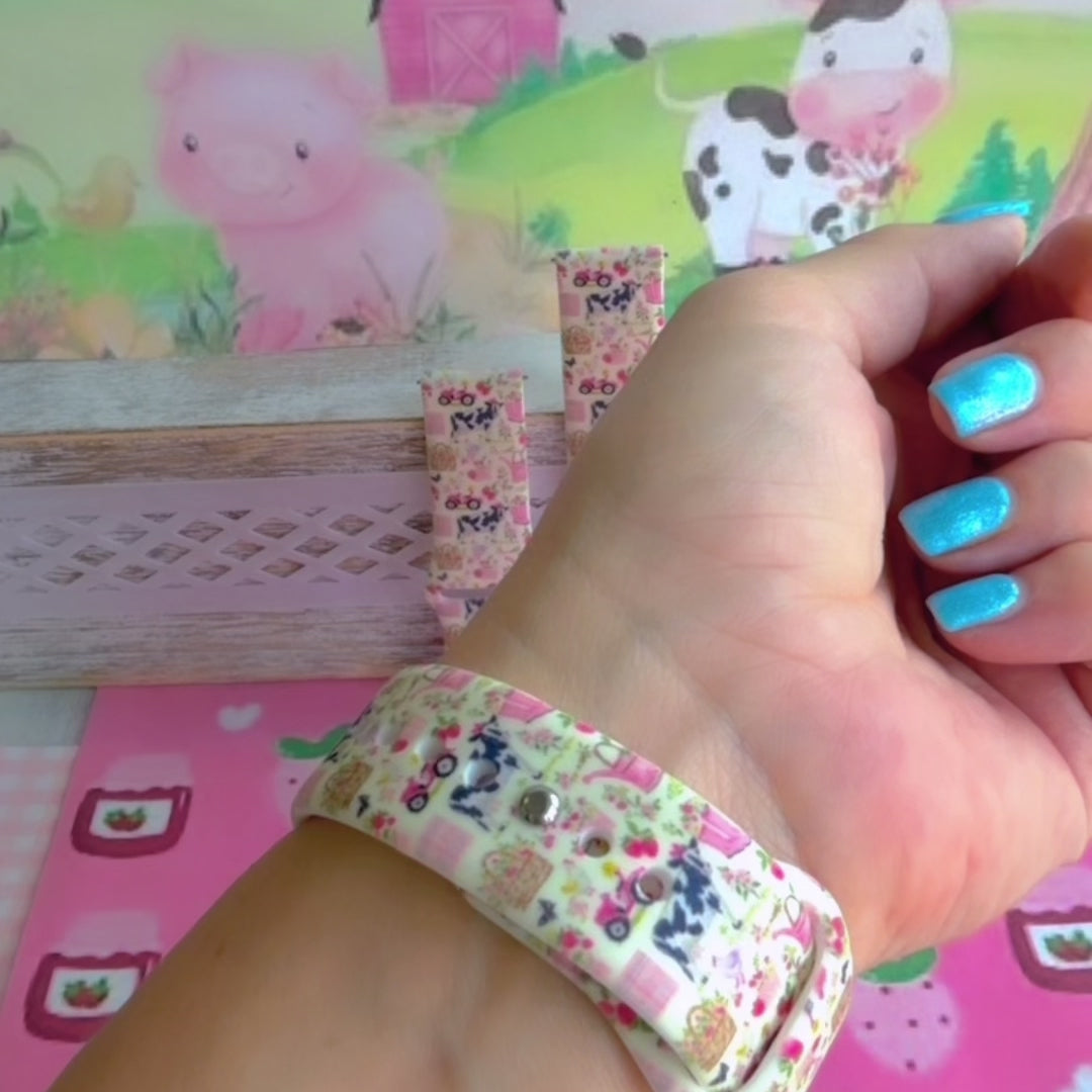 Farm Girl Cutie silicone Samsung Galaxy Watch band with dairy cows, red tractors, chickens, ducks, pink watering cans, strawberries, baskets, and roses on a cream base with pink gingham squares, shown displayed and worn against a farmhouse country backdrop.
