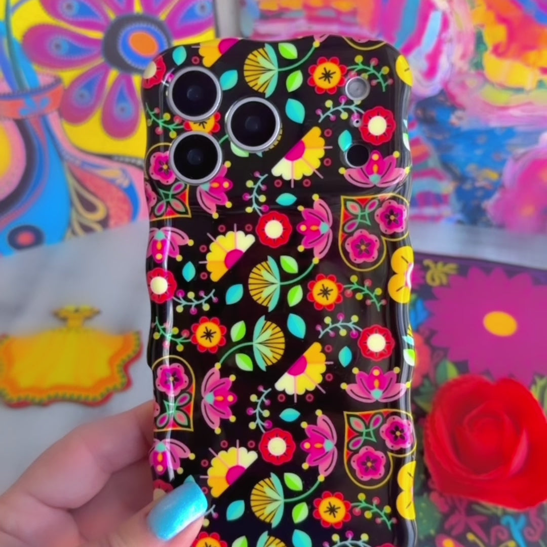 Trendy black wavy iPhone case featuring an intricate folk art botanical pattern of red flowers, magenta details, and yellow fan petals with a high-gloss finish.