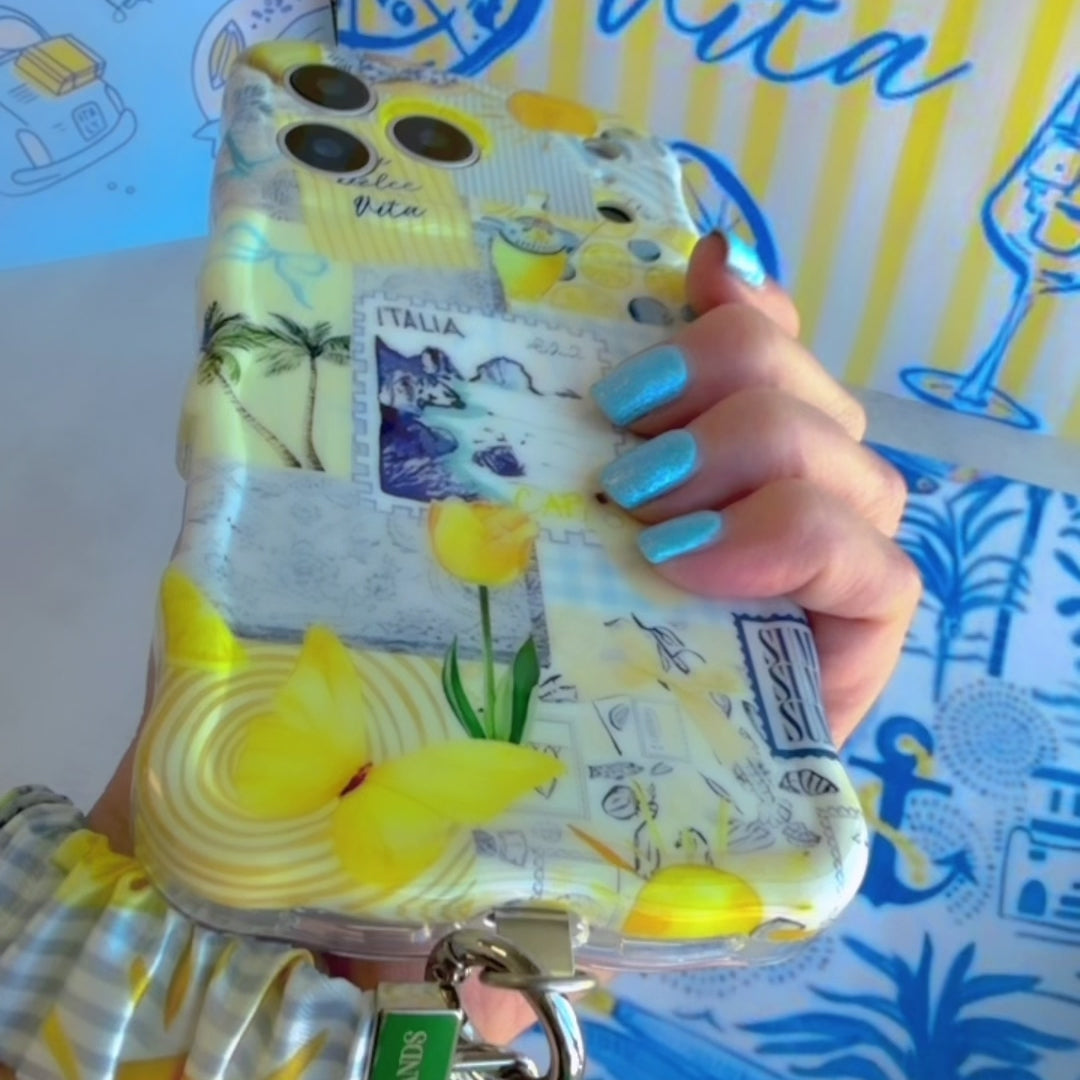 Blue and yellow La Dolce Summer scrunchie phone wrist strap in gingham and lemon print, clipped to the matching Amalfi Coast Wavy Luxe iPhone case with Italia and Capri travel stamps, styled on a blue-and-yellow Mediterranean palm and seashell print background.