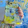 Blue and yellow La Dolce Summer scrunchie phone wrist strap in gingham and lemon print, clipped to the matching Amalfi Coast Wavy Luxe iPhone case with Italia and Capri travel stamps, styled on a blue-and-yellow Mediterranean palm and seashell print background.