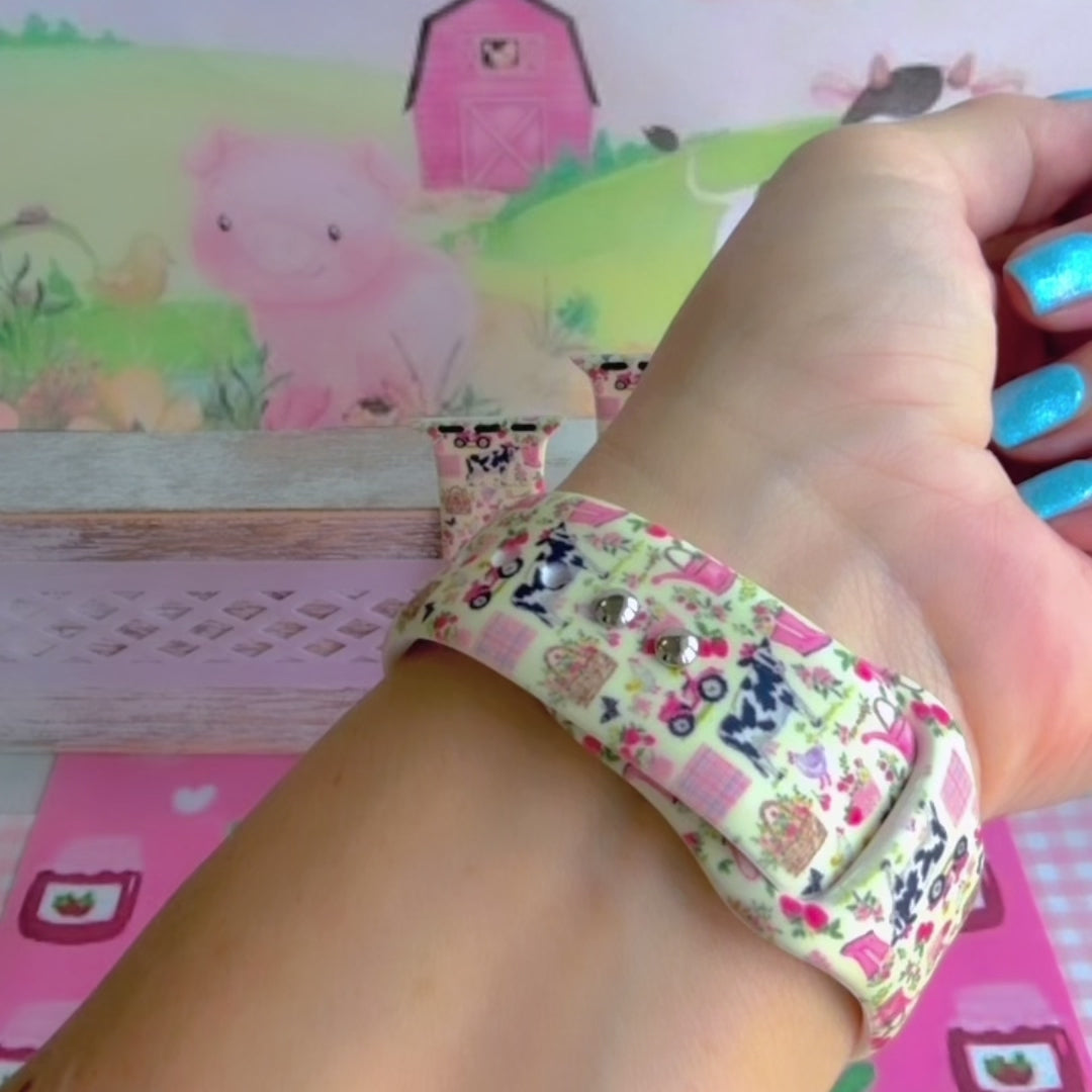 Farm Girl Cutie silicone Apple Watch band with dairy cows, red tractors, chickens, ducks, pink watering cans, strawberries, baskets, and roses on a cream base with pink gingham squares, shown displayed and worn on Apple Watch against a farmhouse country backdrop.
