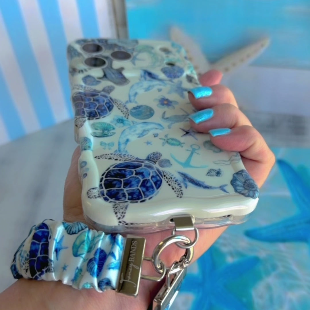 A Blue Lagoon scrunchie phone wrist strap with a blue and white sea turtle and ocean design attached to a matching Wavy Luxe case on a blue starfish background.