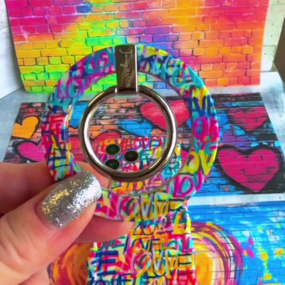 A vibrant Love Riot scrunchie phone wrist strap featuring a colorful neon graffiti "LOVE" and heart design attached to a matching Wavy Luxe phone case on a bright street art background.