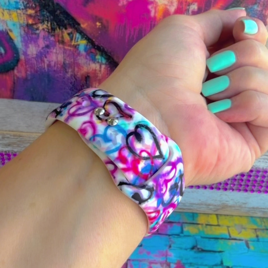 Love Out Loud silicone Apple Watch band with hand-drawn graffiti-style hearts in pink, magenta, blue, teal, red, and black on a white base, shown displayed and worn on Apple Watch against a colorful street art backdrop.