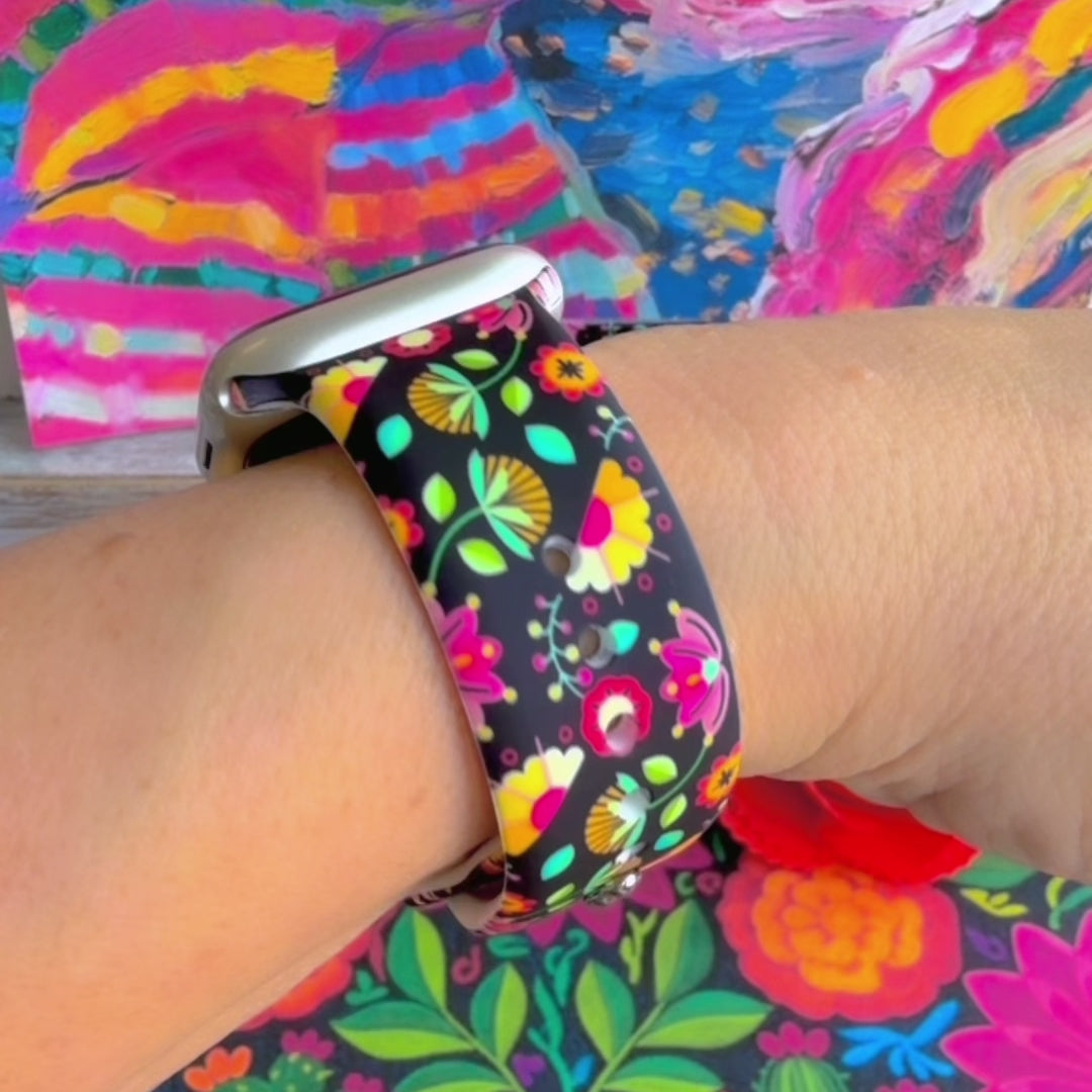 Bloom Bonita silicone Apple Watch band with Mexican folk art florals in coral, red, yellow, teal, and pink on a black base, shown displayed against a vibrant Mexican folk art backdrop with rose and dress accents.