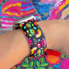 Bloom Bonita silicone Apple Watch band with Mexican folk art florals in coral, red, yellow, teal, and pink on a black base, shown displayed against a vibrant Mexican folk art backdrop with rose and dress accents.