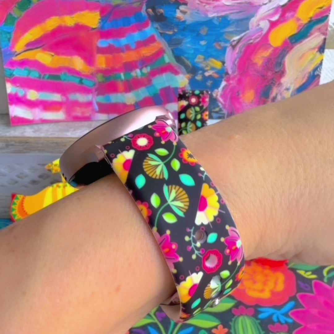 Bloom Bonita silicone Samsung Galaxy Watch band with Mexican folk art florals in coral, red, yellow, teal, and pink on a black base, shown displayed against a vibrant Mexican folk art backdrop with rose and dress accents.