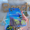 Blue and green satin Beach Please scrunchie phone wrist strap clipped to the matching Beach Please Wavy Luxe iPhone case with a beach chair and cocktail design, styled on a sandy shoreline background with starfish and ocean waves.