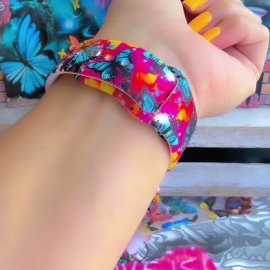 Rebel Wings silicone Samsung Galaxy Watch band with graffiti-style butterflies in blue, orange, yellow, and white over a hot pink and magenta paint-splatter background, shown displayed and worn against a colorful street art butterfly backdrop.