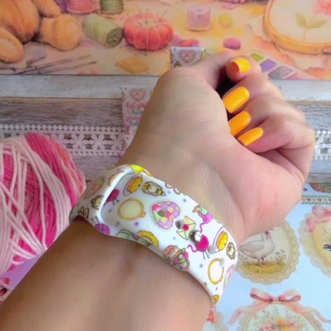 It's Giving Grandma Hobbies silicone Samsung Galaxy Watch band with "hot girls have grandma hobbies" script, patchwork hearts, yarn balls, knitting needles, embroidery hoops, lanterns, pies, and teacups on a cream base, shown displayed and worn against a cottagecore craft backdrop.