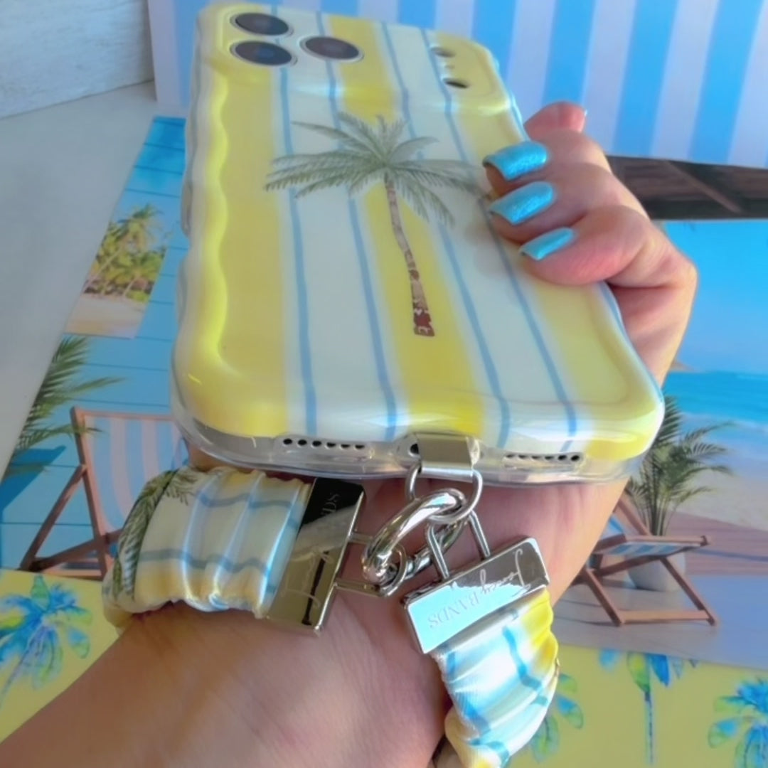 Yellow and blue striped Palm Shore Club scrunchie phone wrist strap with a palm-frond print, clipped to the matching coastal palm Wavy Luxe iPhone case, styled on a sunny yellow beach-club background with blue watercolor palm trees.