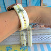 Palm Shore Club silicone Samsung Watch band with hand-painted palm trees over soft blue and yellow cabana stripes on an ivory base, shown displayed and worn on Apple Watch against a coastal beach club backdrop.