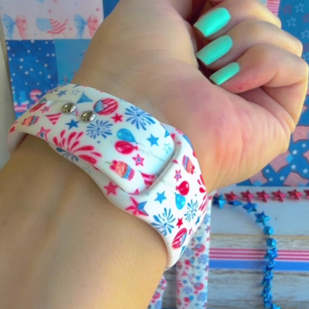 Party In The USA silicone Apple Watch band with fireworks, balloons, cupcakes, flag bunting, and stars in red, white, and blue on a cream base, shown displayed and worn on Apple Watch against a patriotic 4th of July backdrop.