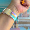 Sweet Summer silicone Samsung Galaxy Watch band with pastel pink, yellow, and blue surfboards, palm trees, and "sweet summer time" script on a soft watercolor beach scene, shown displayed and worn against a coastal surfboard backdrop.