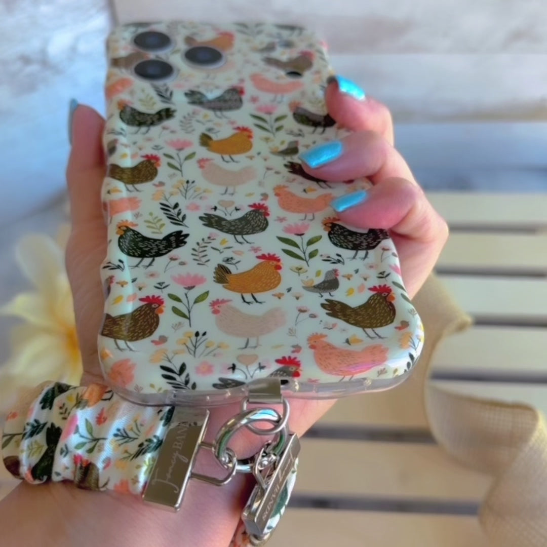 Cream and floral Cluckin' Cute scrunchie phone wrist strap with a hand-illustrated chicken and wildflower print, clipped to the matching farmhouse chicken Wavy Luxe iPhone case, styled on a rustic wood and burlap background.