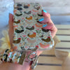 Cream and floral Cluckin' Cute scrunchie phone wrist strap with a hand-illustrated chicken and wildflower print, clipped to the matching farmhouse chicken Wavy Luxe iPhone case, styled on a rustic wood and burlap background.