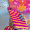 Pink and orange striped Candy Citrus Stripes scrunchie phone wrist strap clipped to the matching hot pink and citrus orange striped Wavy Luxe iPhone case, styled on a playful pink and orange heart print background.